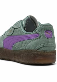 Baskets en suède vert avec une surface texturée, dotées d'une bande latérale violette et d'une semelle en caoutchouc marron. Comprend un petit logo au niveau du talon.