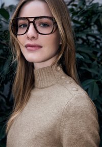 Brauner Strickpullover mit hohem Kragen und Knopfdetails an der Schulter, kombiniert mit großen Hornbrillen.