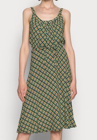 Grön blommig klänning med tunna axelband, figursydd vid midjan, med ett upprepande mönster i orange och gult, lätt tyg och svängig nederdel.