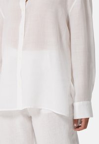 Camicia button-up bianca in lino con vestibilità rilassata, caratterizzata da una leggera trasparenza, maniche lunghe e dettagli con patta dei bottoni.