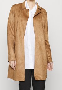 Brun mockaskinnsjacka med krage, långa ärmar och öppen framsida. Kombinerad med en vit skjorta med knappar och svarta byxor. Slät yta.