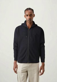 Muž oblečený v čiernom zipsovom mikinovom svetri s šnúrkami a béžových cargo nohaviciach, stojaci na pozadí s jednoduchým svetlým vzorom.