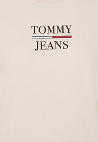 Krämfärgad tyg med texten "TOMMY JEANS" i svart, åtskild av ett litet horisontellt streck i vitt, rött och svart.