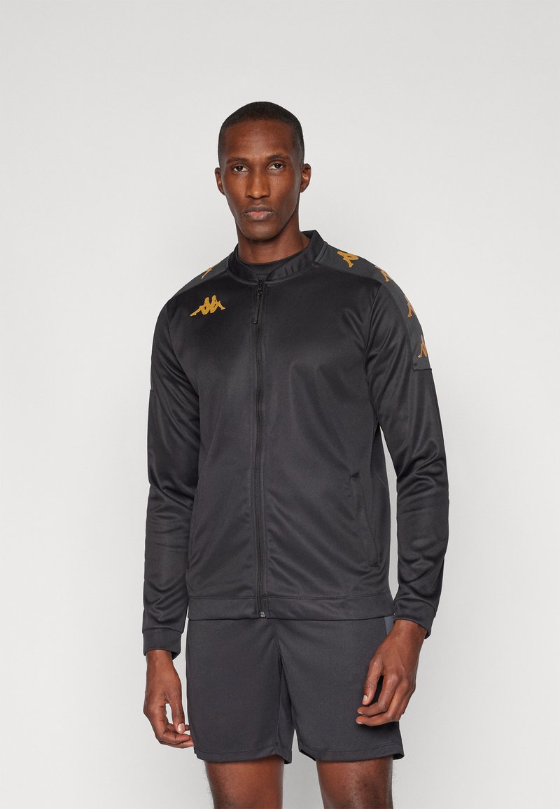 Veste de sport noire en tissu lisse avec fermeture éclair, ornée de détails dorés et d'un logo sur les épaules. Portée avec un short assorti.
