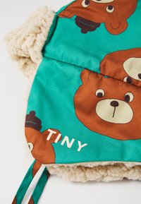 Tissu turquoise avec des motifs d'ours brun, présentant une doublure en beige pelucheux et le mot "TINY" en blanc. Deux bretelles à nouer ajustables.