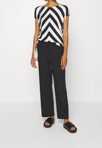 Frau trägt ein schwarz-weiß gestreiftes Oberteil im Chevron-Muster, schwarze weit geschnittene Hosen, schwarze Slippers und eine kleine beigefarbene Umhängetasche.