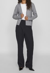 Grå stickad cardigan med V-hals och ribbade manschetter, bärs över en vit skjorta. Matchas med svarta vida byxor och svarta klackar.