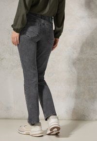 Jeans in denim grigio con una vestibilità slim, cuciture visibili e due tasche posteriori. Indossati con sneakers beige e una maglietta larga a maniche lunghe color oliva.