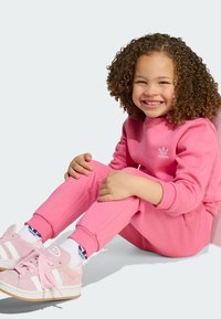 Ružová mikina a zladené teplákové nohavice vyrobené z mäkkého materiálu. Výrazné biele logo Adidas na hrudi. Ružové topánky s bielymi pruhmi a šnúrkami.