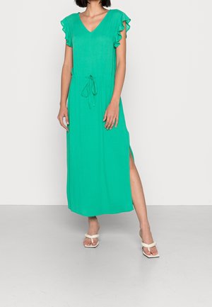 Vestito verde di media lunghezza con maniche corte arricciate, scollo a V, vita con coulisse e spacchi laterali. Abbinato a sandali bianchi.