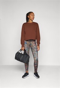Sweatshirt court marron, leggings à motif coloré et baskets noires. Tenu d'un sac de sport noir, debout devant un fond neutre.