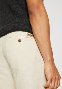 Pantaloni beige in tessuto strutturato, con un bottone marrone visibile e un'etichetta in pelle INDICODE sulla vita. Maglione scuro sopra.
