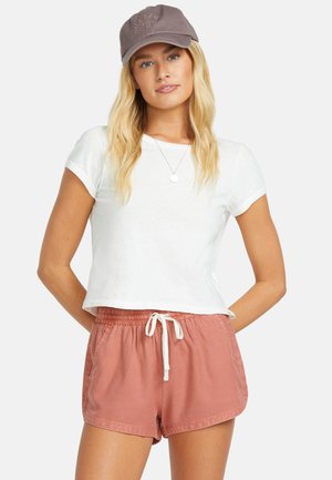 Blondynka w szarej czapce z daszkiem, białym krótkim topie odsłaniającym brzuch, złotym naszyjniku z zawieszką oraz rdzawego koloru szortach z troczkiem, stojąca z rękami za plecami.