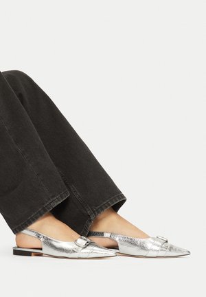 Nohy v stříbrných špičatých balerínách na podpatku se sponami, spojené s černými rovnými kalhotami na jednoduchém světlém pozadí.