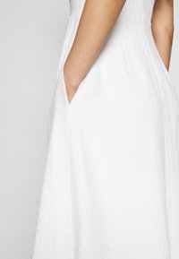 Bras placé dans la poche latérale d'une robe blanche texturée sans manches avec une silhouette en A.