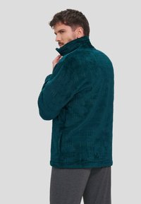 Chaqueta de forro polar color verde azulado con cuello alto, patrón de cuadrícula texturizado, bolsillos laterales y un corte holgado. Lleva encima de pantalones grises.