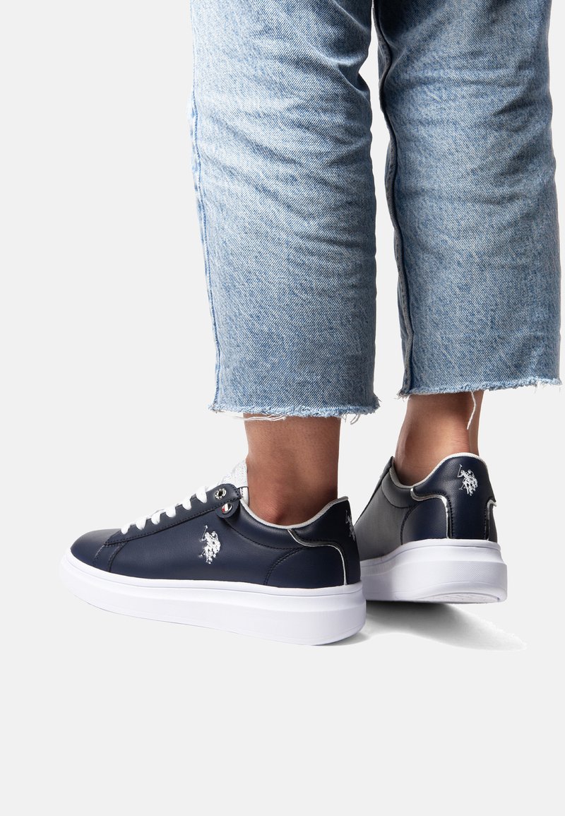 Navyblauwe leren sneakers met witte zolen, witte veters en een logodetail. Beschikt over een gladde textuur en afgeronde vorm, met versleten denim jeans.