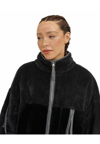 Giacca in pile nera con colletto alto, chiusura con zip e due tasche con zip. Caratterizzata da un corpo testurizzato con accenti grigi lisci.