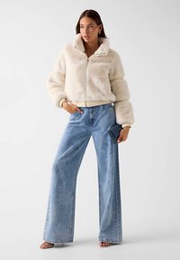 Veste zippée en fausse fourrure crème avec un col montant, associée à un jean à jambes larges bleu clair. Le modèle tient une pochette bleu foncé.