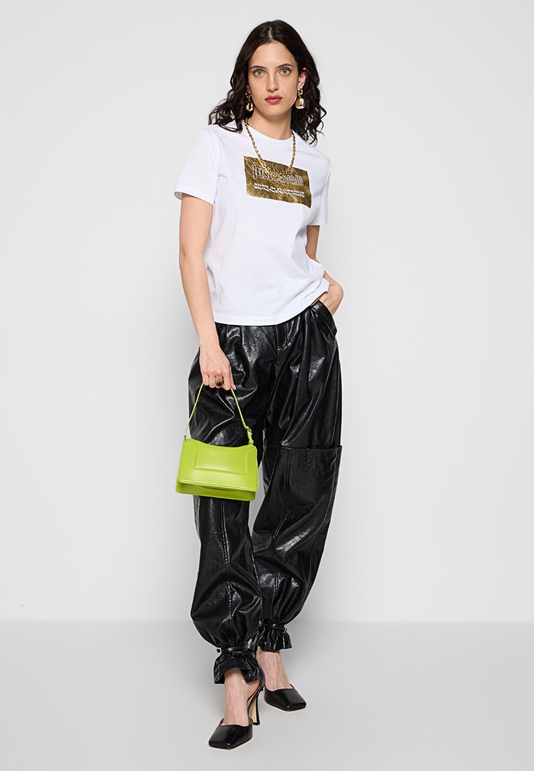 Mujer con camiseta gráfica blanca, pantalones de cuero negros brillantes con tobillos recogidos, tacones negros, joyería dorada y sosteniendo un pequeño bolso verde lima.