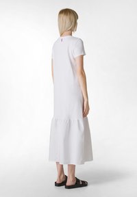 Vestido de algodón blanco con mangas cortas, un corte holgado, dobladillo en capas y un pequeño acento rosa en la parte posterior. Llevado con sandalias negras.