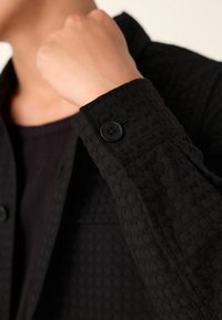 Schwarze Texturjacke mit einem gemusterten Stoff und einem Knopf am Ärmel, die ein klassisches und strukturiertes Design zeigt.