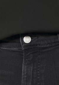 Vaqueros de mezclilla negra con un botón plateado que lleva la inscripción "Tommy Jeans New York". Primer plano de la cintura que muestra una costura fina y la textura.