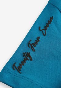 Camiseta de tela turquesa con texto en cursiva negra "Veinticuatro Siete" en la manga. Textura suave y ajuste casual.