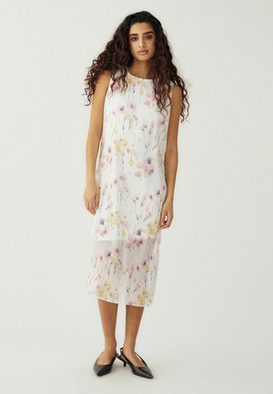 Žena s dlhými kučeravými vlasmi má na sebe biele midi šaty bez rukávov s ružovo-žltým kvetinovým vzorom a čierne slingback lodičky, stojaca na bielom pozadí.