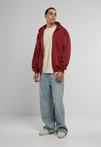 Maroon zip-up hoodie with a kangaroo pocket, paired with a light cream t-shirt and loose light blue jeans. Sneakers complete the outfit.