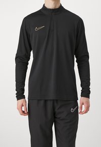 Schwarzes langärmliges Sportoberteil mit einem Reißverschlusskragen; verfügt über ein goldenes Nike-Logo auf der Brust. Glattes Material und enganliegendes Design.