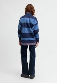 Svītrots džemperis ar ziliem un tumši ziliem horizontāliem svītrām, pavasara audums, brīvs piegriezums, augsta apkakle, pāri tam uzvilkts raibais krekls, kombinēts ar izplestiem džinsiem.