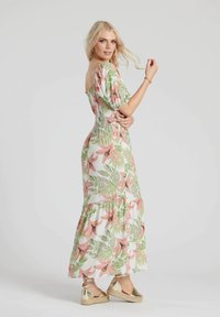 Blommig maxiklänning med vit bakgrund, med rosa liljor och gröna blad. Smockad liv och flerlagd nederdel; bärs med guldig espadrillos.