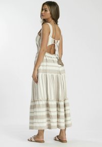 Vestido maxi en una tela ligera con rayas beige, que presenta una espalda baja, un lazo ajustable y un dobladillo en capas. Combinado con sandalias planas.