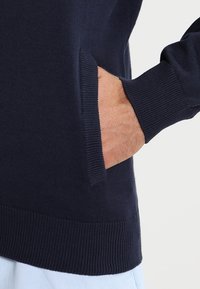 Tmavě modrý svetr s žebrovaným výstřihem a manžetami, vybavený boční kapsou. Vyrobený z měkkého pleteného materiálu, zdůrazňujícího texturu a pohodlí.