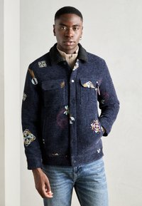 Ναυτικό μπλε σακάκι με λουλουδάτα σχέδια, δύο τσέπες στο στήθος και μαύρο γούνινο γιακά. Φορεμένο πάνω από ένα μπεζ τοπ, συνδυασμένο με ανοιχτό μπλε τζιν.