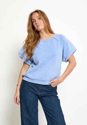 Vrouw met lang blond haar, gekleed in een lichtblauw topje met korte mouwen en donkerblauwe jeans, staand met één hand in haar zak.