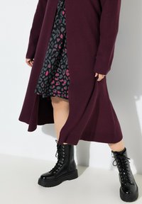 Burgundy neulecardigan kerroksittain mustan mekon päällä, jossa on vaaleanpunaisia ja harmaita leopardikuoseja, täydentäen asua mustat nauhakengät, joissa on paksut pohjat.