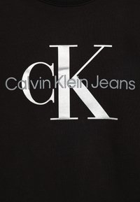 Felpa nera con grandi lettere bianche e argento che scrivono "Calvin Klein Jeans" con "CK" mostrato in modo prominente in una tipografia stilizzata.