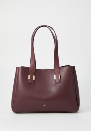 Sac fourre-tout en cuir marron foncé avec deux poignées et de petites décorations rectangulaires en métal doré, posé debout sur une surface blanche.