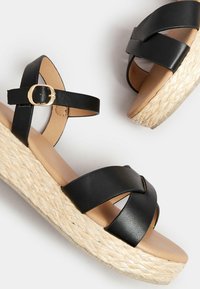 Mustat nahka wedgesandaalit, joissa on ristiin menevät remmit, juuttikäyttäinen alusta ja kultainen solki. Sileä pinta ja avoin varvasmuoto.