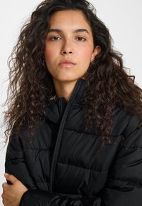 Vrouw met lang krullend donker haar, gekleed in een zwarte gewatteerde jas, die recht vooruit kijkt met haar armen over elkaar geslagen.