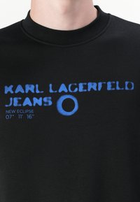 Sudadera de algodón negra con "KARL LAGERFELD JEANS" en texto azul en negrita, con un acento circular y la fecha "07' 11' 16" impresa debajo.