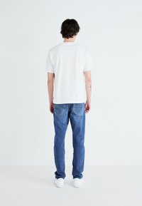 Bela kratka majica s kratkimi rokavi, kombinirana z modrimi jeans hlačami in belimi športnimi čevlji, prikazana s hrbta. Gladek material in oprijet kroj z ojačanimi šivi.