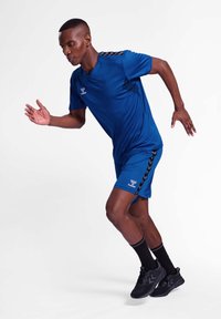 Blå atletisk skjorte og shorts med sorte sidelænder, sat mod en hvid baggrund. Modellen er i en løbestilling, iført sorte sko og sokker.