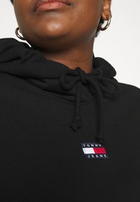 Sudadera negra con cordón, con un parche rectangular del logo de Tommy Jeans en azul, rojo y blanco. Textura de tela suave, diseño casual.