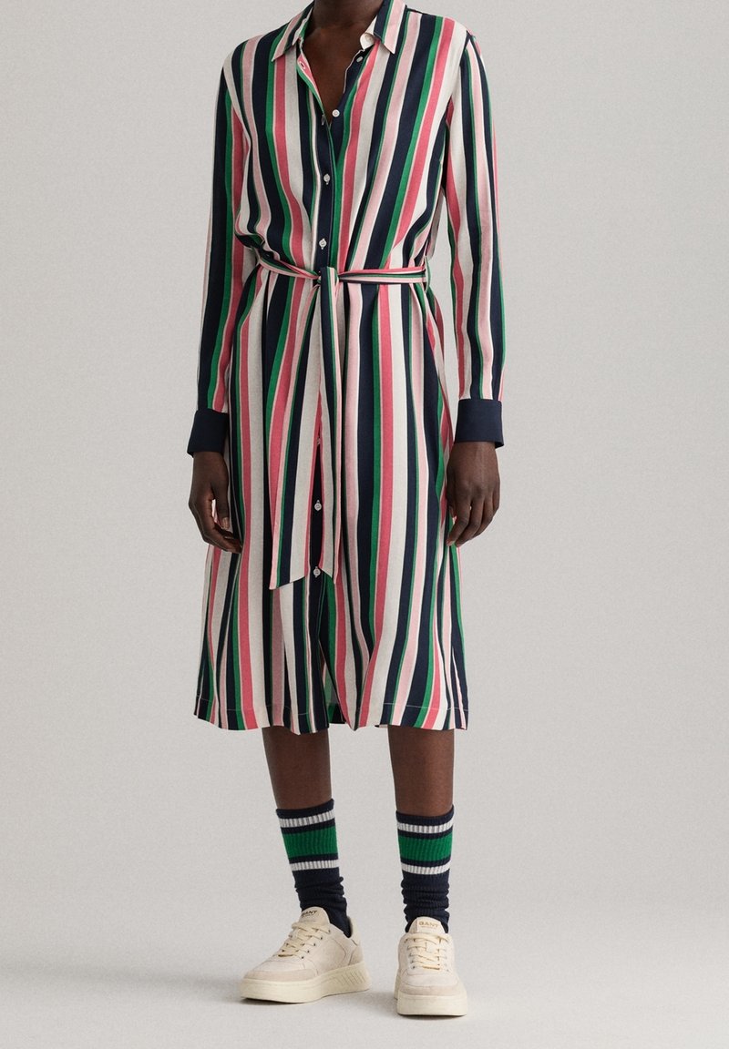 Robe chemise rayée en rose, vert et marine, faite d'un tissu léger, avec des manches longues, un système de fermeture par boutons et une ceinture à la taille. Associée à des baskets et des chaussettes rayées.