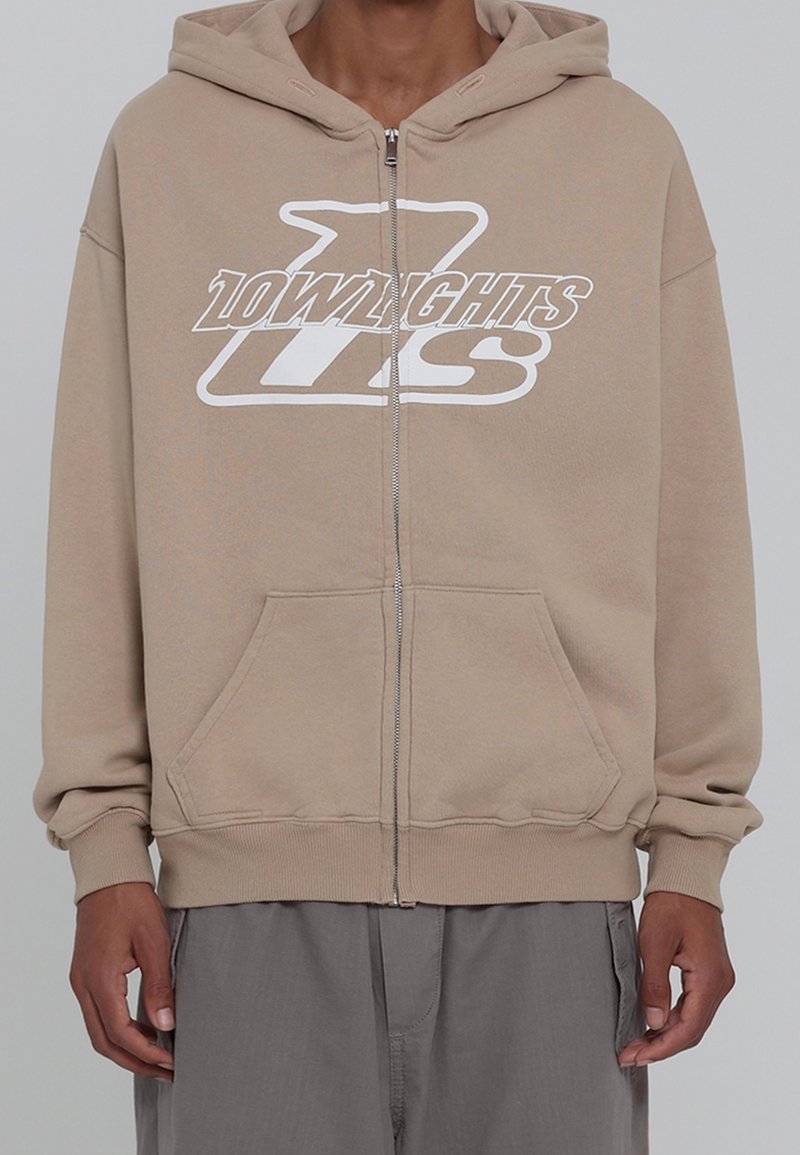 Personne portant un sweat à capuche beige zippé avec le logo "10LOWLIGHTS" et "TLS" en blanc, associé à un pantalon gris ample.