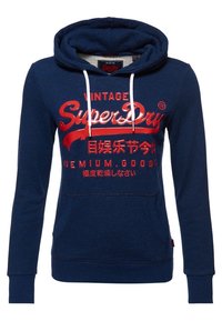 Sudadera azul marino con cordones blancos, bolsillo frontal y logo rojo de "SuperDry" junto con texto en japonés impreso en el pecho.