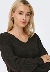 Svart, strukturstrikket genser med bred V-hals og lange ermer. Modellen har blondt hår og sølvfargede hoops.